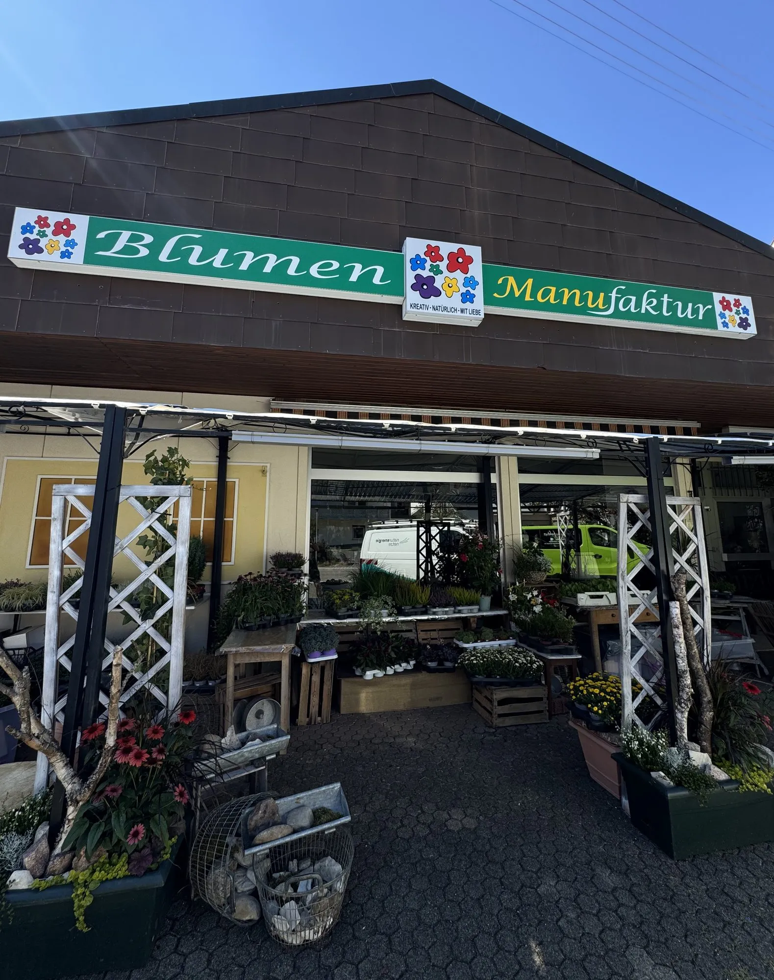 Die Blumen Manufaktur von außen: Ladenschild, Vordach und die Pflanzenauslage auf dem Pflaster